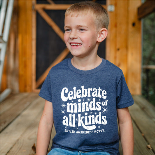 Child wearing a blue t-shirt with 'Celebrate minds of all kinds' text, sitting on a wooden floor.