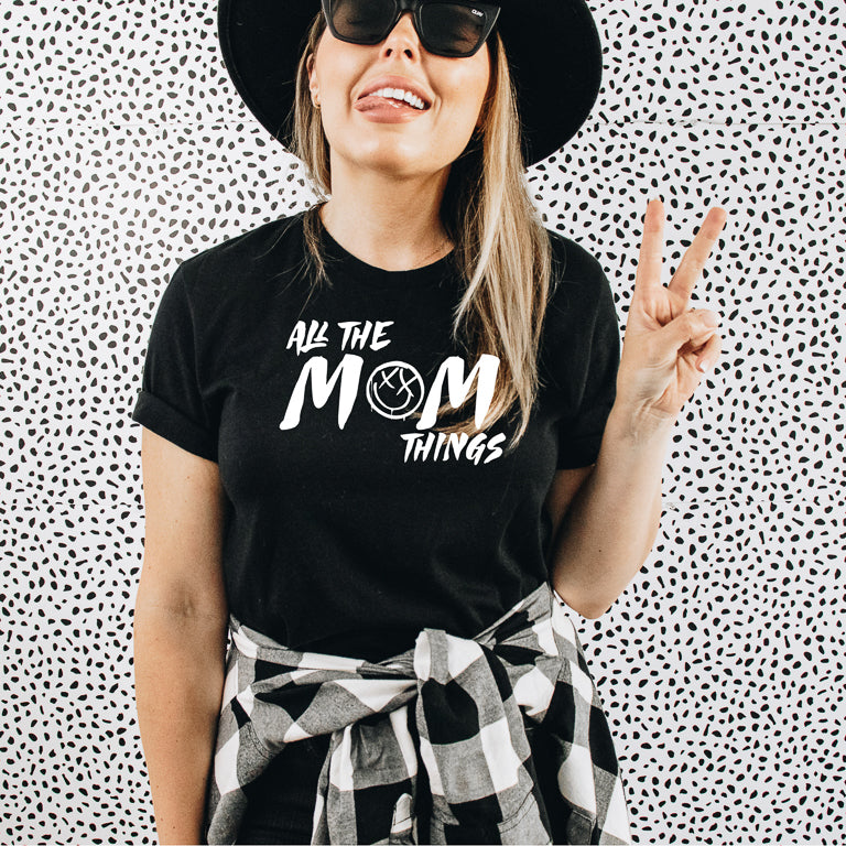 Woman wearing a black t-shirt with 'All the Mom Things' text, making a peace sign against a patterned background.