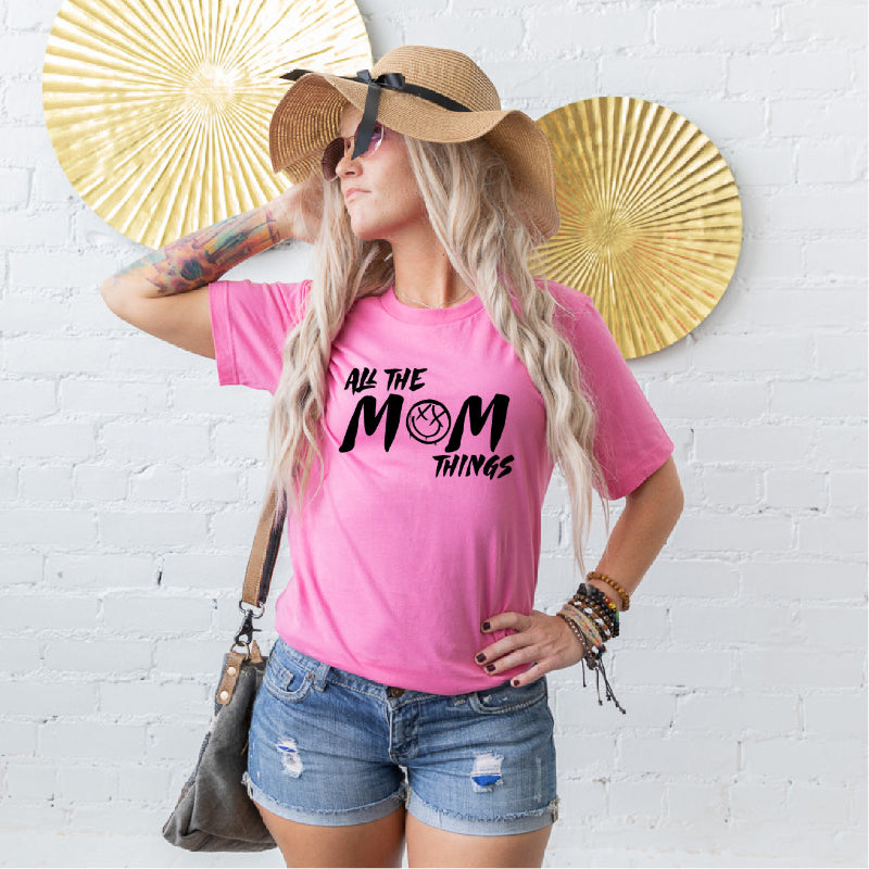 Woman wearing a pink t-shirt with 'All the Mom Things' text, standing against a white brick wall with gold decorative elements.