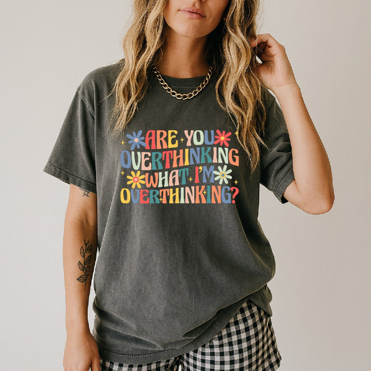 A woman wearing a dark gray acid-wash graphic t-shirt with a colorful retro text design that reads 'ARE YOU OVERTHINKING WHAT I'M OVERTHINKING?'. She is also wearing a gold chain necklace and a black and white gingham patterned skirt, photographed against a neutral light gray background.
