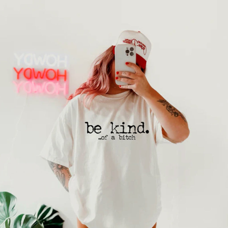 Person wearing a white t-shirt with 'be kind. . .of a bitch' text, holding a phone with a colorful case.