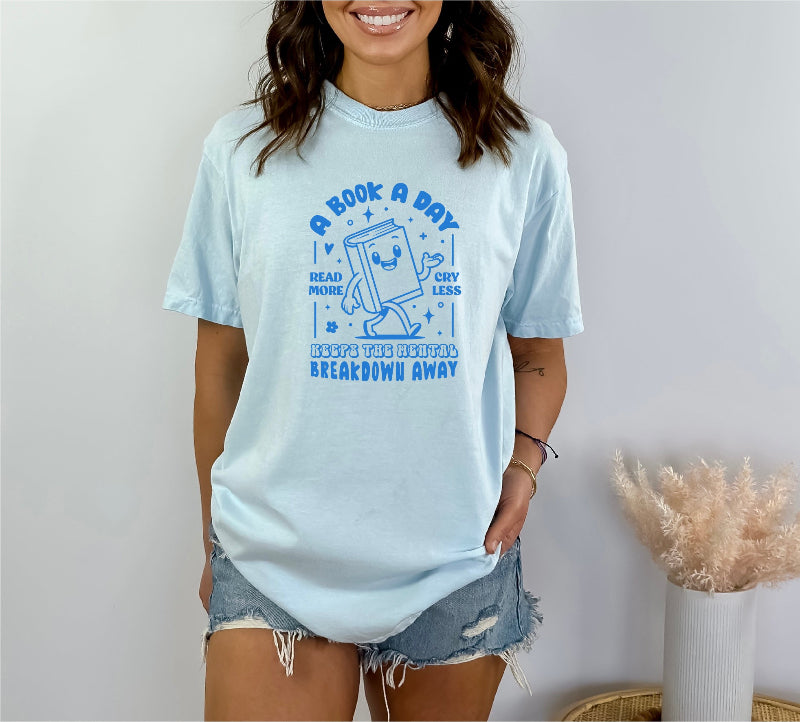 a book a day keeps the mental breakdown away dark blue on light blue unisex tee pictured on a woman with jean shorts. 
