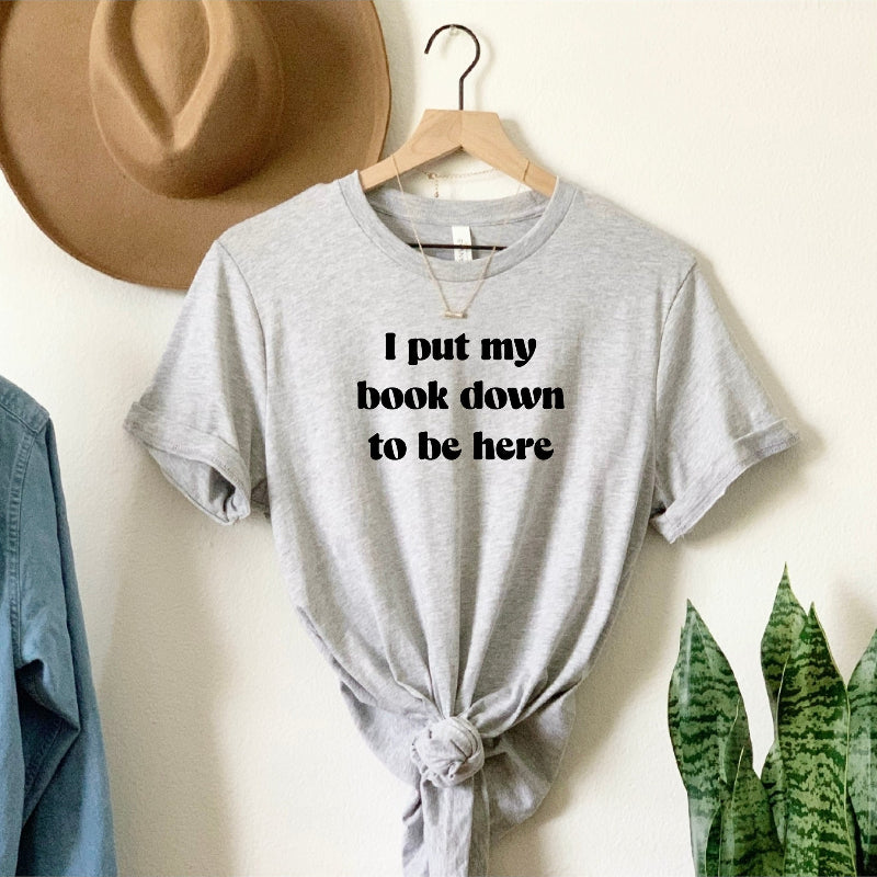 Gray t-shirt with text 'I put my book down to be here' on a hanger against a neutral wall.