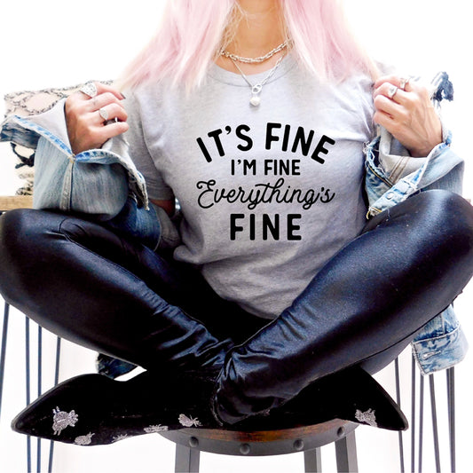 woman with pink hair sitting cross legged on stool with grey shirt that reads its fine im fine everything's fine