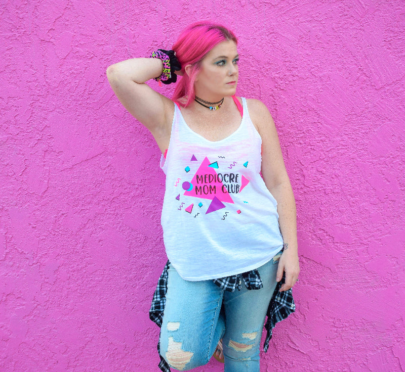 Person wearing a white tank top with colorful text that reads "mediocre mom club" and graphics against a pink wall.