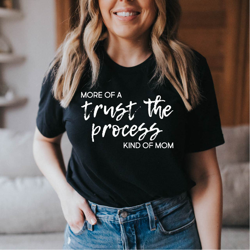 Woman wearing a black t-shirt with text that reads "more of a trust the process kind of mom", standing in a casual indoor setting.