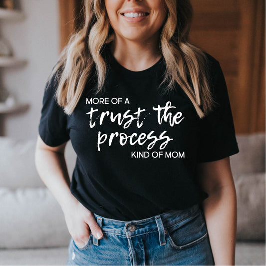 Woman wearing a black t-shirt with text that reads "more of a trust the process kind of mom", standing in a casual indoor setting.