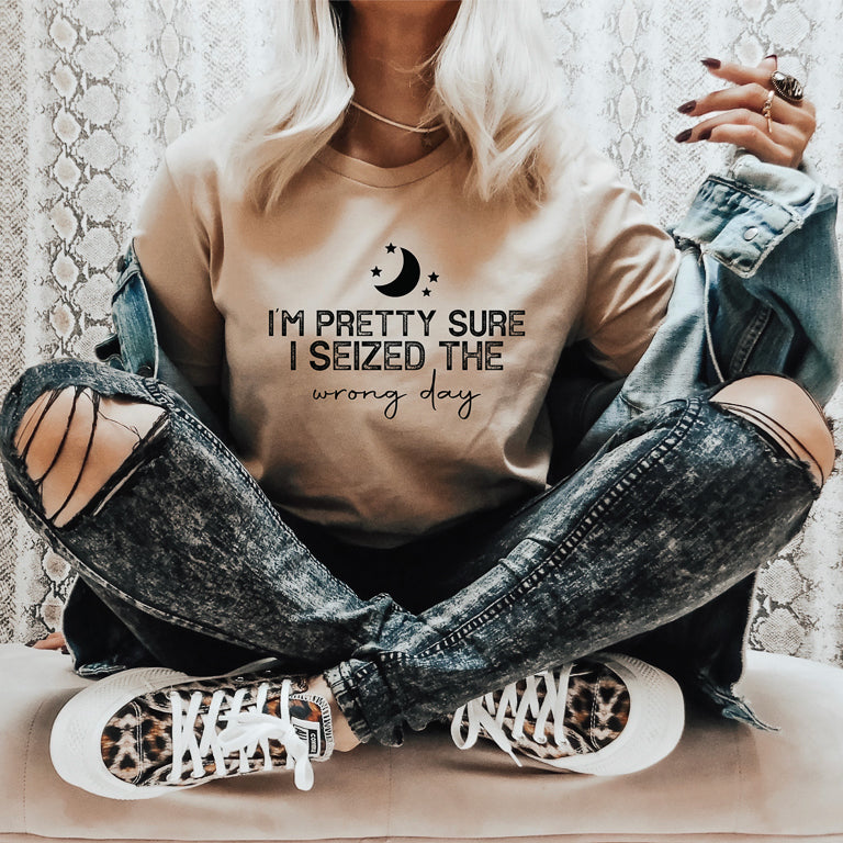 Person wearing a beige t-shirt with text that reads pretty sure I seized the wrong day and ripped jeans sitting on a surface.