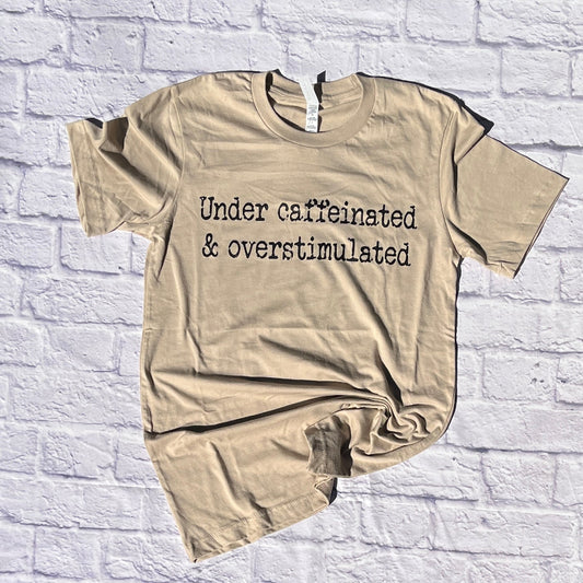 tan tshirt that reads under caffeinated and overstimulated on white brick background